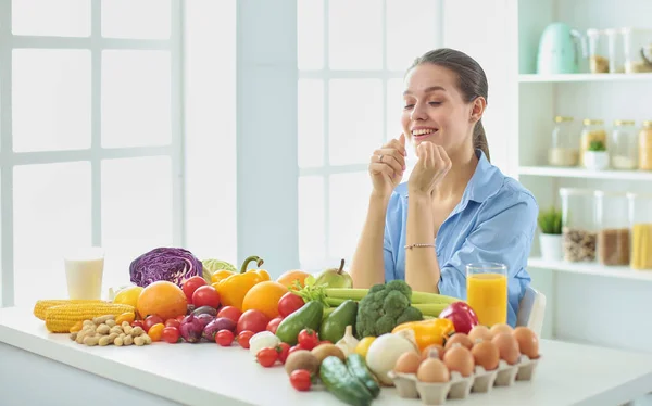 Happy young housewife sitting in the kitchen preparing food from a pile of diverse fresh organic fruits and vegetables, selective focus — Stock Photo, Image