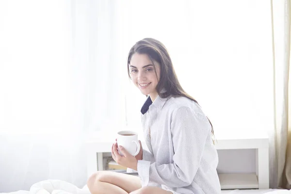 Mladá krásná šťastná brunetka žena v černý hedvábný župan ležel v posteli, pít ranní kávu, bílý šálek. Dámské dovolené den — Stock fotografie