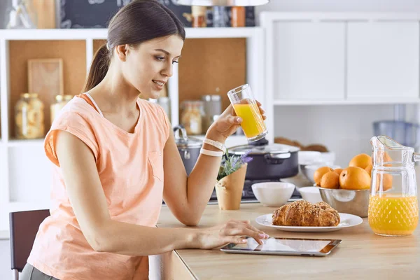 Mujer bonita sonriente mirando el teléfono móvil y sosteniendo un vaso de jugo de naranja mientras desayunaba en una cocina — Foto de Stock