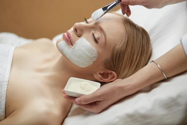 people, beauty, cosmetology and treatment concept - close up of beautiful young woman lying with closed eyes and cosmetologist applying facial mask by brush at spa