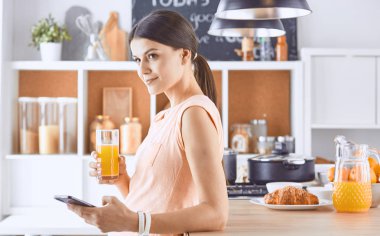 Cep telefonu görünümlü ve cam portakal suyu bir mutfakta Kahvaltı yaparken tutan gülümseyen güzel kadın