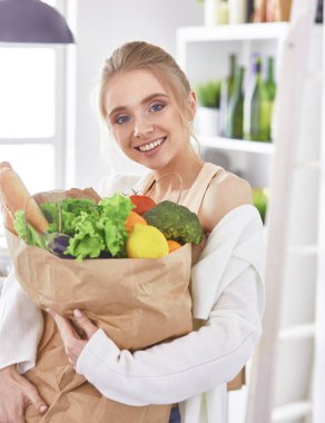 Sebzeli alışveriş poşeti taşıyan genç bir kadın mutfakta duruyor.