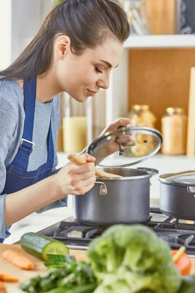 Hermosa chica está degustando comida y sonriendo mientras cocina en casa — Foto de Stock