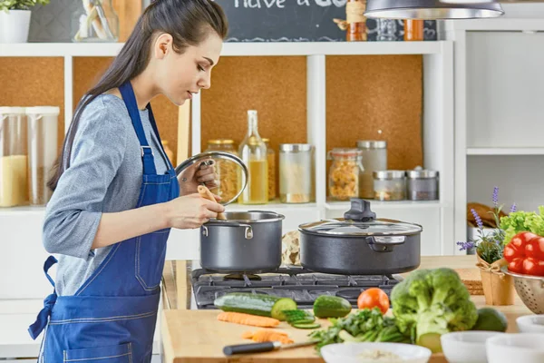 Bella ragazza sta assaggiando il cibo e sorridendo mentre cucina in cucina a casa — Foto Stock