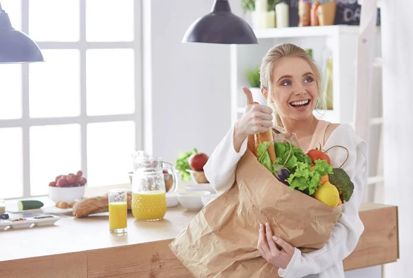 Молода жінка тримає продуктовий сумку з овочами стоїть на кухні — стокове фото