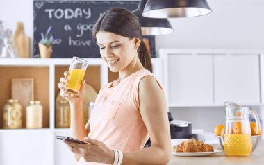 Cep telefonu görünümlü ve cam portakal suyu bir mutfakta Kahvaltı yaparken tutan gülümseyen güzel kadın