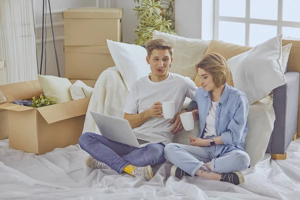 Retrato de pareja feliz mirando el ordenador portátil juntos sentados en una casa nueva, rodeados de cajas — Foto de Stock