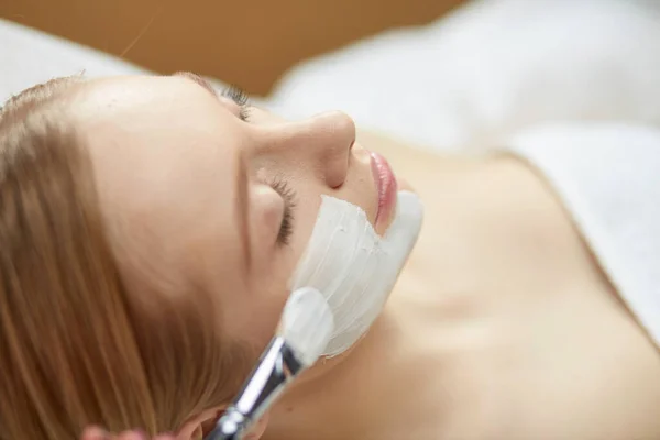 Mensen, schoonheid, visagie en behandeling concept - close-up van de mooie jonge vrouw liggen met gesloten ogen en schoonheidsspecialist gezichts masker toe te passen door borstel in spa — Stockfoto