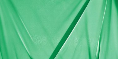 Yakın plan parlak yeşil kumaş. Görünür dokuları olan saf ve hafif bir malzeme. Kumaş örtülmüş ve katlanmış. Görüntü derinliği ve hareket hissi veren soyut, dalgalı şekiller ve gölgeler yaratıyor.