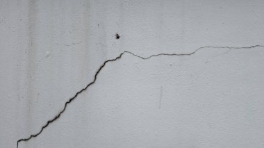 Beyaz ya da açık gri beton bir duvarın yakın çekimi ve belirgin siyah bir çatlak. Betonun pürüzlü, desenli yüzeyi açıkça görülüyor. Kırığın ince koyu çizgisi çerçeve boyunca çapraz çalışır