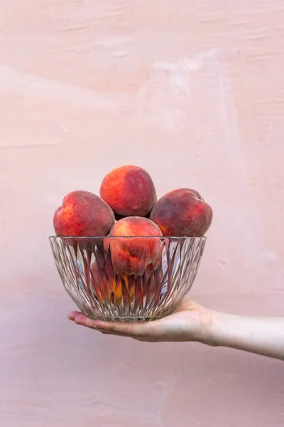 Close up vertical photo of woman hand hold ripe mellow peaches in large glass crystal transparent bowl isolated on pastel light bright background