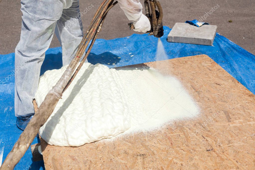 Close up view of technician dressed in a protective white uniform spraying foam insulation using Plural Component Spray Gun. Spraying polyurethane foam for roof and energy saving