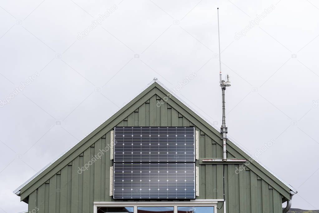 Cropped photo of little green house with small solar panel at the front of the building next to the cellular antenna against gray blue sky
