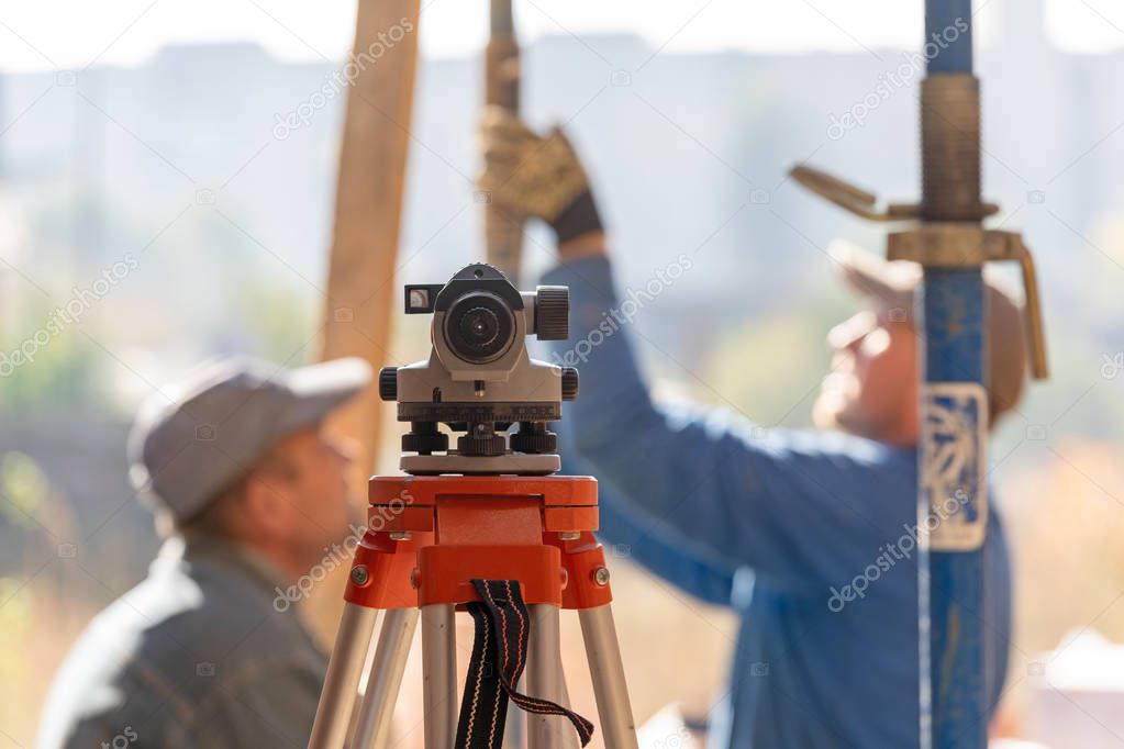 Formwork construction concept. Photo of optical level device against blurred background with two adult unrecognizable professional workman