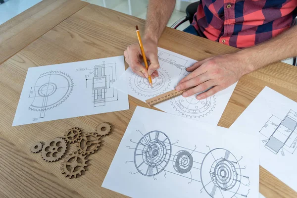 明るいインテリアの木製の机の後ろに座って静かな男のトリミング 彼はいくつかの歯車要素と描画技術面に定規と鉛筆を使用してください — ストック写真