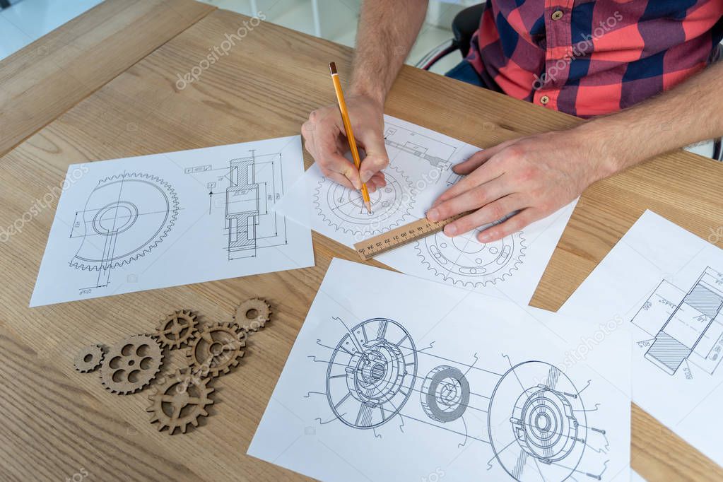 Cropped photo of calm man sitting behind wooden desk in bright interior. He using pencil and ruler to making technical drawing with few cogwheel elements
