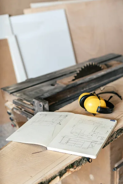 Vertical photo of notebook with technical drawing lying on top of wood plank board in garage or workroom near yellow headset and table saw