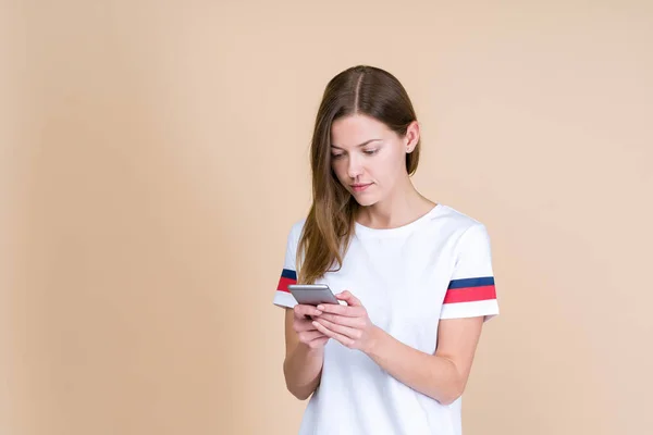 Mujer joven de pie sobre fondo beige pastel con teléfono inteligente —  Fotos de Stock