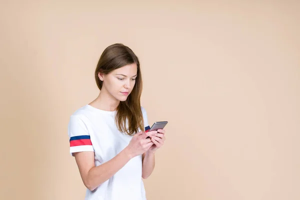 Mladá žena stojící na pastelové béžové pozadí s chytrým telefonem — Stock fotografie