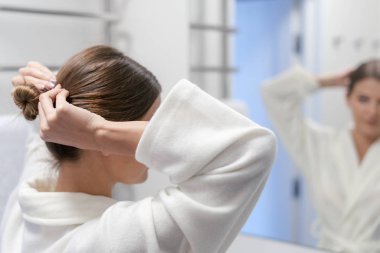 Beyaz bornozlu kadın banyodaki aynaya bakıyor.