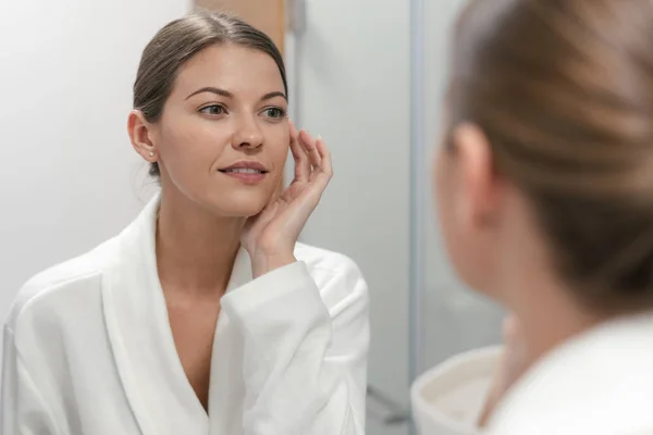 Mujer en albornoz mirando en el espejo en el baño — Foto de Stock