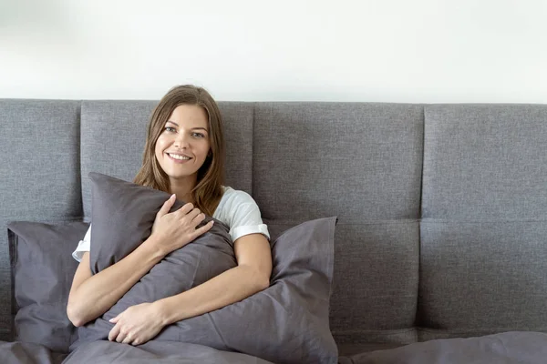 Junge erwachsene Frau sitzt auf Bett und umarmt Kissen — Stockfoto