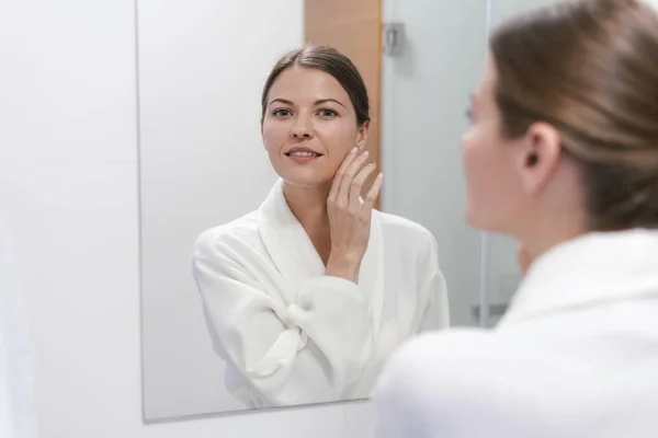 Woman in bathrobe looking in mirror at bathroom — Stock Photo, Image
