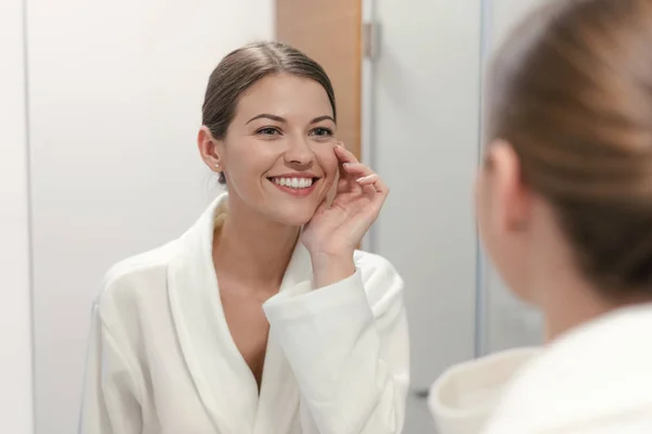 Mujer en albornoz mirando en el espejo en el baño — Foto de Stock