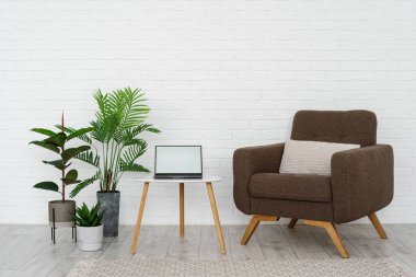 Minimalist oturma odası minimalist mobilya, rahat koltuk, ahşap üçgen sehpa, halı, dizüstü bilgisayar ve beyaz tuğla duvara karşı yeşil bitkiler sergileniyor.