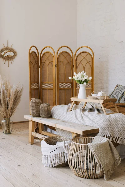 Vertical photo of comfortable bedroom in bohemian interior style with breakfast tray flowers and textile sheet on bed wooden bench seat bamboo dressing screen home decor in wicker basket Stock Image