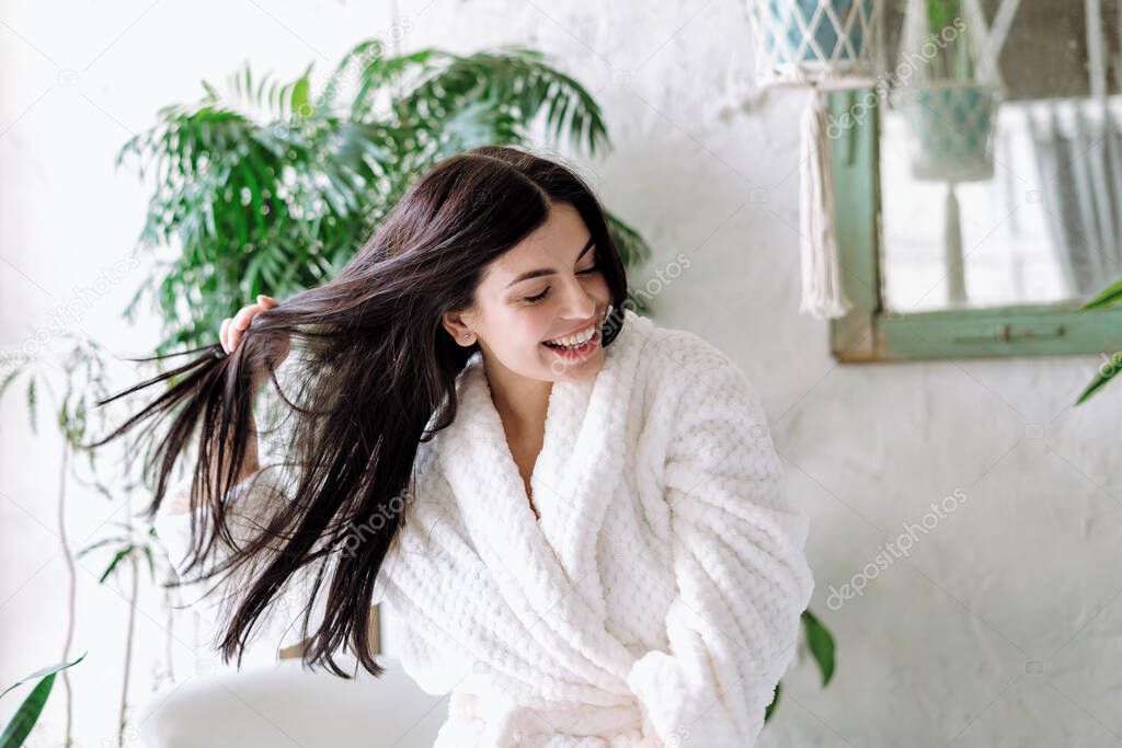 Smiling woman shaking head with floating hair, laughing wide, having fun at home. Happy young adult female in bathrobe spending morning at bathroom