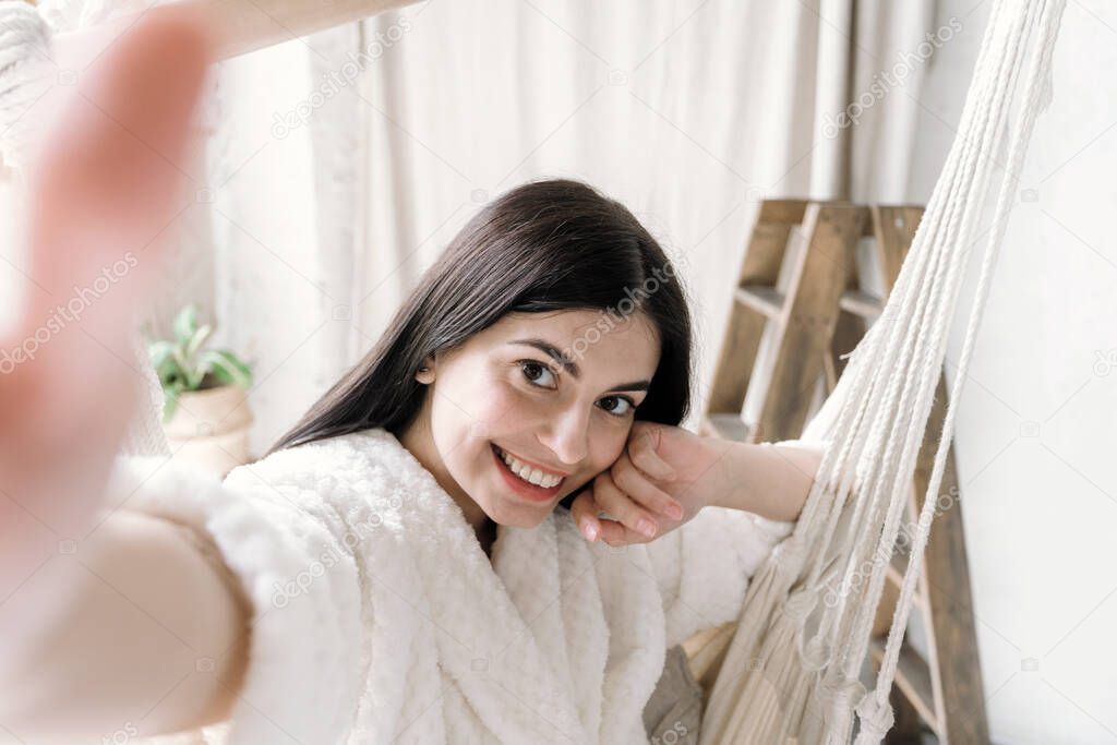 Portrait view of happy young adult woman in bathrobe sitting on a hanging swing, making self photo on camera, smiling wide and having fun, resting at home on weekend