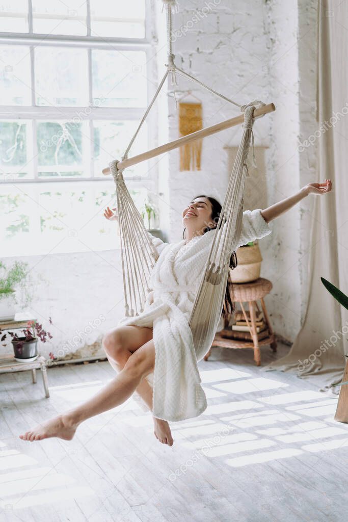 Vertical photo of carefree young adult female in bathrobe sitting on a hanging swing at home, enjoying lazy weekend morning, having fun and raised hands up, feeling free