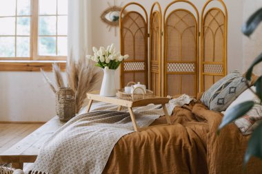 Laleler ve kahvaltı tepsisi çaydanlık ve fincanla yatakta. Bohem tarzında bir apartman dairesi Hygge yatak odası, ahşap bank koltuğu, bambu giyinme perdesi, ev dekorasyonu, vazoda kuru bitkiler.
