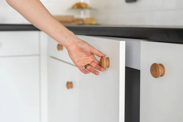 Cropped view of woman pulling wooden handle, open white door on cupboards standing at contemporary kitchen. Element of drawers under countertop in modern house