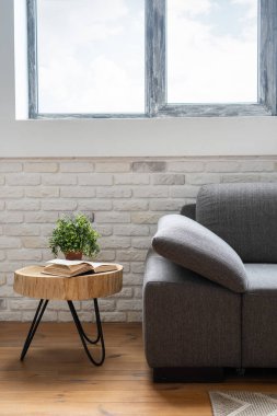 Pencerenin altındaki gri tekstil kanepenin dikey görünümü çağdaş iç kısımda yeni mobilyalarla birlikte. Çiçek saksısında kitap ve bitki, sehpanın üstünde, ahşap zeminin yanında.