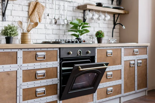 Modern built in oven with open door. Side view of wooden kitchen in contemporary interior house with new furniture, gas stove equipment, kitchenware supplies and houseplant in flower pot