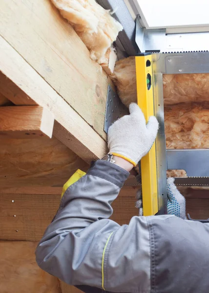 Concept of house improvement. Vertical and cropped view of professional worker man using measuring level, checking surface near new mansard skylight window, standing indoor building under construction