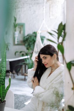 Bornozlu genç bir kadının dikey profili, sabahları banyoda, sağlıklı esmer saçlara bakarak ve güzel gülümseyerek geçirmesi.