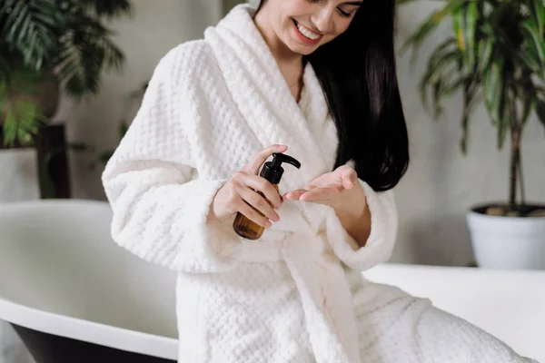 快乐的年轻成年女性穿着浴衣 早上在浴室里 用天然保湿乳液的玻璃瓶 妇女洗澡时 应用护肤化妆品 — 图库照片