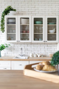 Lavaboya, musluğa, mutfak gereçlerine, dolaptaki mobilyalara ev bitkilerine, masadaki bulanık kahvaltıya seçici bir şekilde odaklan. Modern evdeki beyaz iç döşemeli mutfağın dikey görüntüsü