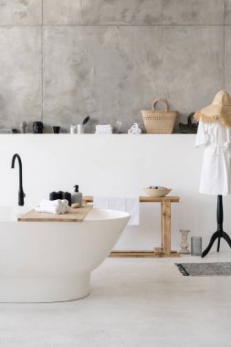 Modern banyo içerisindeki beyaz çağdaş evde dikey fotoğraf boş küvette ahşap raflarda havlular, bornoz ve arka planda ev dekorasyonu.