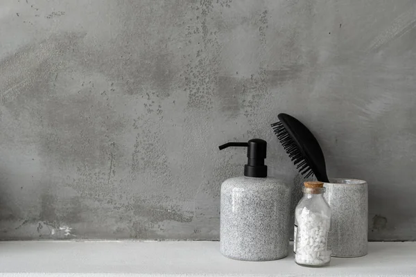 Beauty objects against grey copy space wall in bathroom. Close up of bottle with soap dispenser, hair comb and bath salt in room with modern interior design