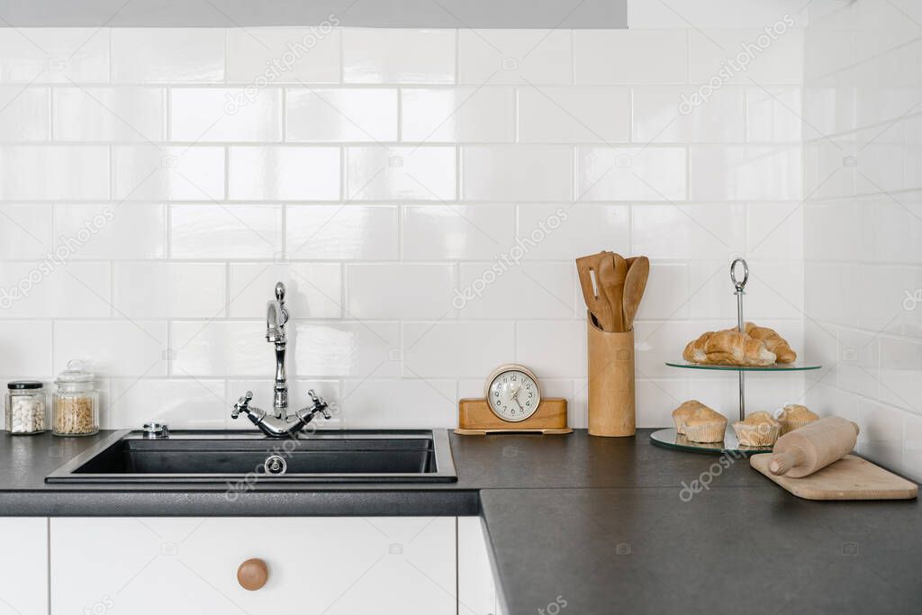 White kitchen in modern house with sink, water tap, utensils on black countertop over drawers against copy space tiled wall