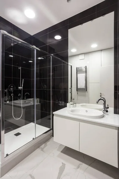 Vertical view of modern house with contemporary interior design in bathroom, shower cabin, sink on countertop, mirror and heated towel rail against black tiled wall