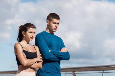 Dışarıda spor eğitimi yapan iki ciddi arkadaşın yan görüntüsü. Yan yana durup ellerini göğsünün üstüne koyup yan yana duruyorlar. Sporcu ve sporcu kadının düşük açılı görüntüsü