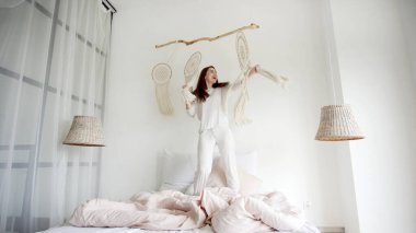 Hafta sonu sabah konsepti. Genç ve mutlu bir kadının rahat yatak odasında pijamalarıyla dans edişinin geniş bir manzarası var. Yatakta duruyor, sırıtıyor ve şarkı söylüyor.