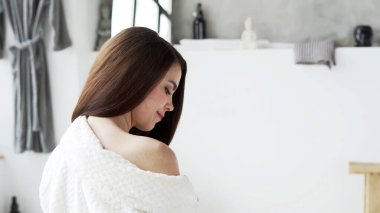 Güzellik konsepti. Beyaz bornozlu, gülümseyen genç bir kadının profil görüntüsü. Sabahı banyoda, yan tarafta oturarak evde fotokopi alanının arka planında oturuyor.