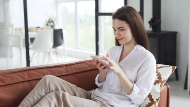 Kanepede oturan, modern akıllı telefonu kullanan, yeni uygulamadan bilgi kontrol eden, internette sörf yapan ve hafta sonları evde dinlenen sıradan kıyafetlerle gülümseyen genç bir kadının yan görüntüsü.