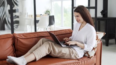 Uzaktan çalışma konsepti. Kanepede oturan, modern dizüstü bilgisayar kullanan, evdeki online işleri kontrol eden gündelik giysili mutlu genç bir kadın.